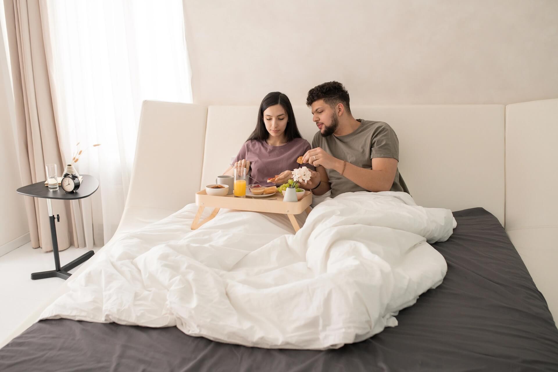 Young amorous couple in t shirts sitting on bed having breakfast